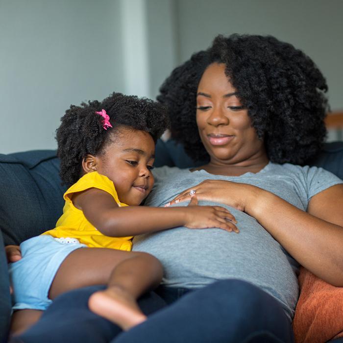 A pregnant Black woman with natural hair is reclining on a dark blue couch, holding her toddler daughter. They are both looking at and smiling at the woman's pregnant belly.