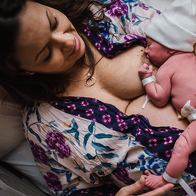 A woman is reclining, wearing a robe with printed flowers, she is breastfeeding a newborn infant who's wearing a knit cap