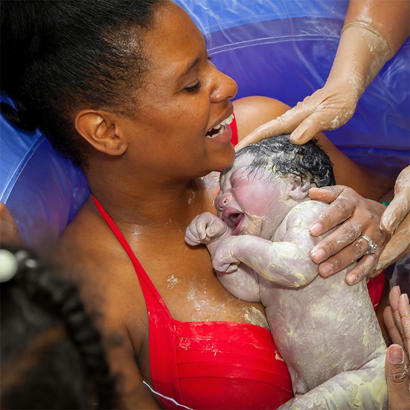 A happy Black woman in a water birth setting is being handed her infant who is covered in vernix caseosa